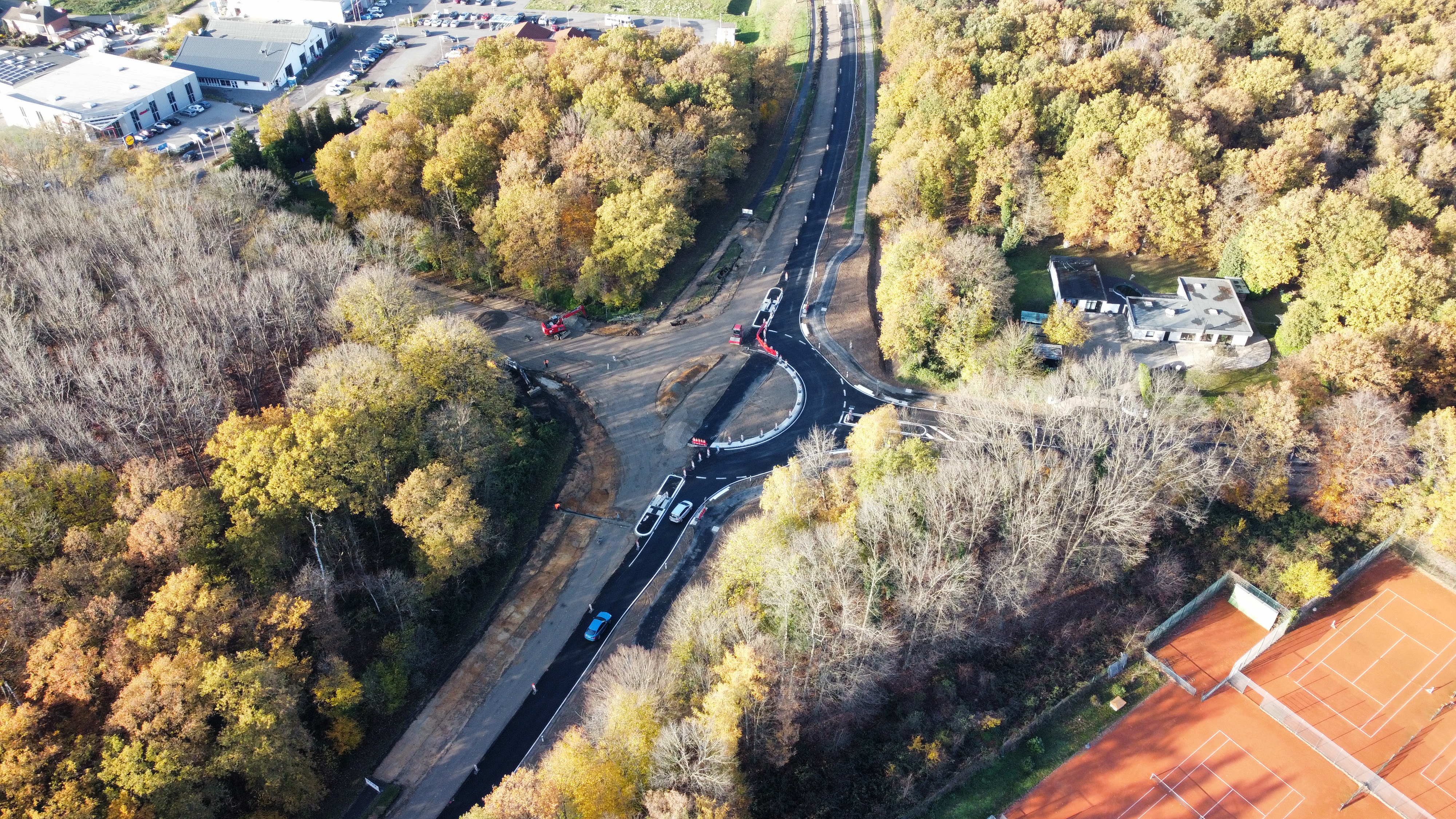 Luftbild von Baustelle für Neubau Kreisverkehrkreuzung B221/Borner Straße/Boisheimer Straße mit fertiggestellter rechten Kreisverkehrseite und Fahrbahn umgeben von dichtem, verschiedenartigen Baumbewuchs vereinzelt Autos auf der Straße, rechte Ecke unten Ausschnitt vom Tennisplatz, linke obere Ecke Ausschnitt von Einkaufsmeile auf der Borner Straße mit Fitnessstudio und Restaurant